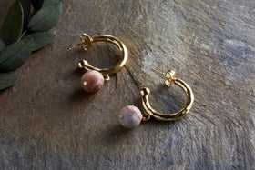 The Cerita Rhodochrosite Earrings - Rhodochrosite & Gold by nkuku rest elegantly on textured stone, with green leaves in the corner adding a natural touch to this stylish jewellery gift display.