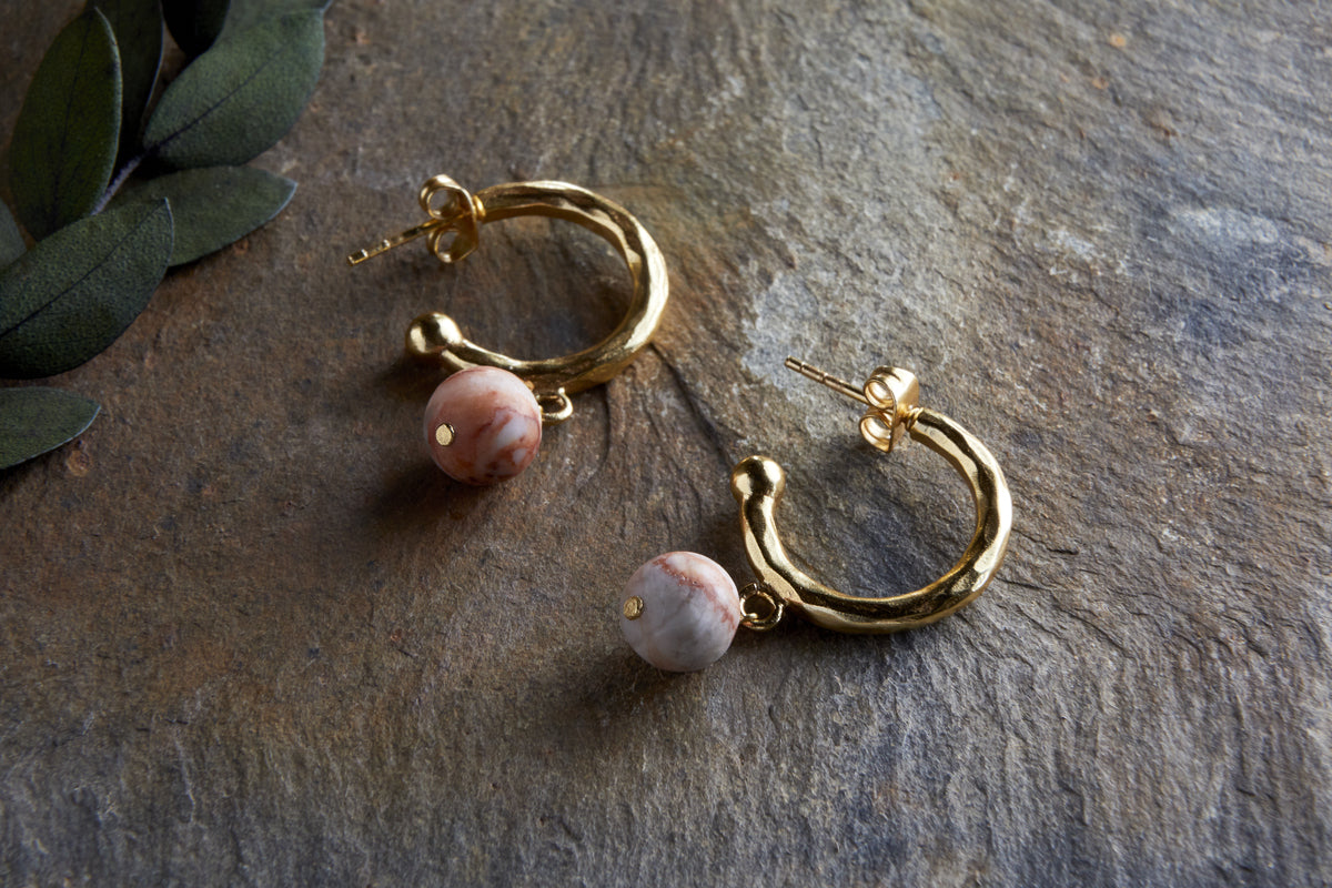 The Cerita Rhodochrosite Earrings - Rhodochrosite & Gold by nkuku rest elegantly on textured stone, with green leaves in the corner adding a natural touch to this stylish jewellery gift display.