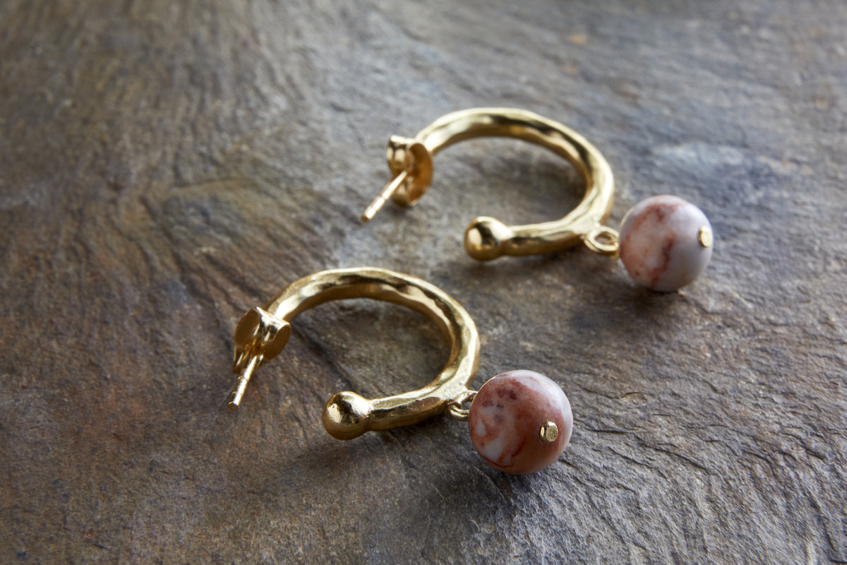 The nkuku Cerita Rhodochrosite Earrings feature gold-plated hoops with a textured finish, each adorned with a round rhodochrosite bead—an elegant jewellery gift for any occasion.