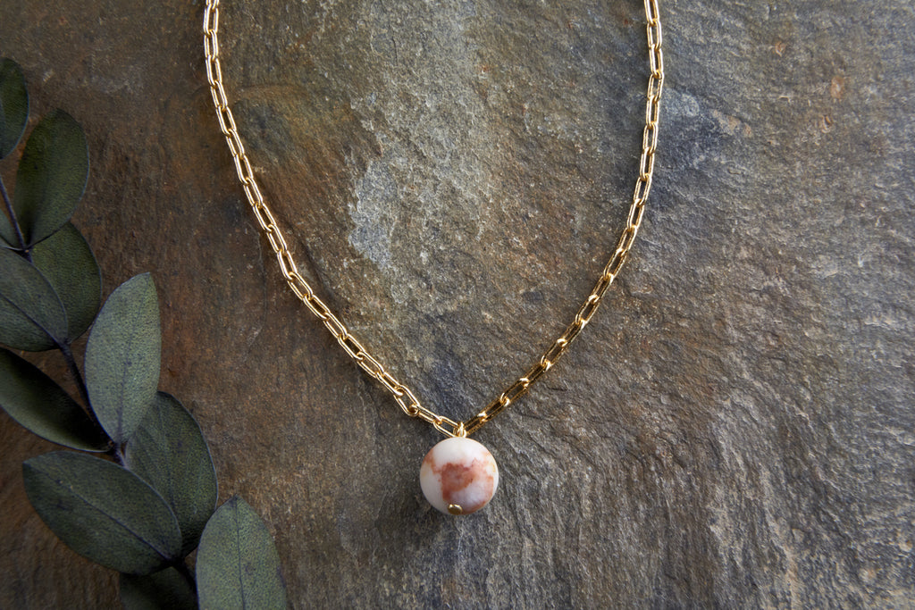 The nkuku Cerita Rhodochrosite Necklace features a marbled pink and white rhodochrosite pendant on an elongated gold chain. Displayed on textured gray stone with three green leaves, it makes an elegant jewelry gift.