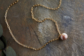 The Cerita Rhodochrosite Necklace by nkuku features elongated gold links and a gold-plated pendant set with a round, marbled rhodochrosite stone, displayed on a textured gray stone surface with green leaves nearby.