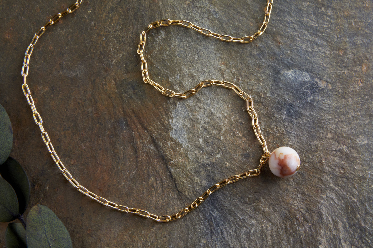 The Cerita Rhodochrosite Necklace by nkuku features elongated gold links and a gold-plated pendant set with a round, marbled rhodochrosite stone, displayed on a textured gray stone surface with green leaves nearby.