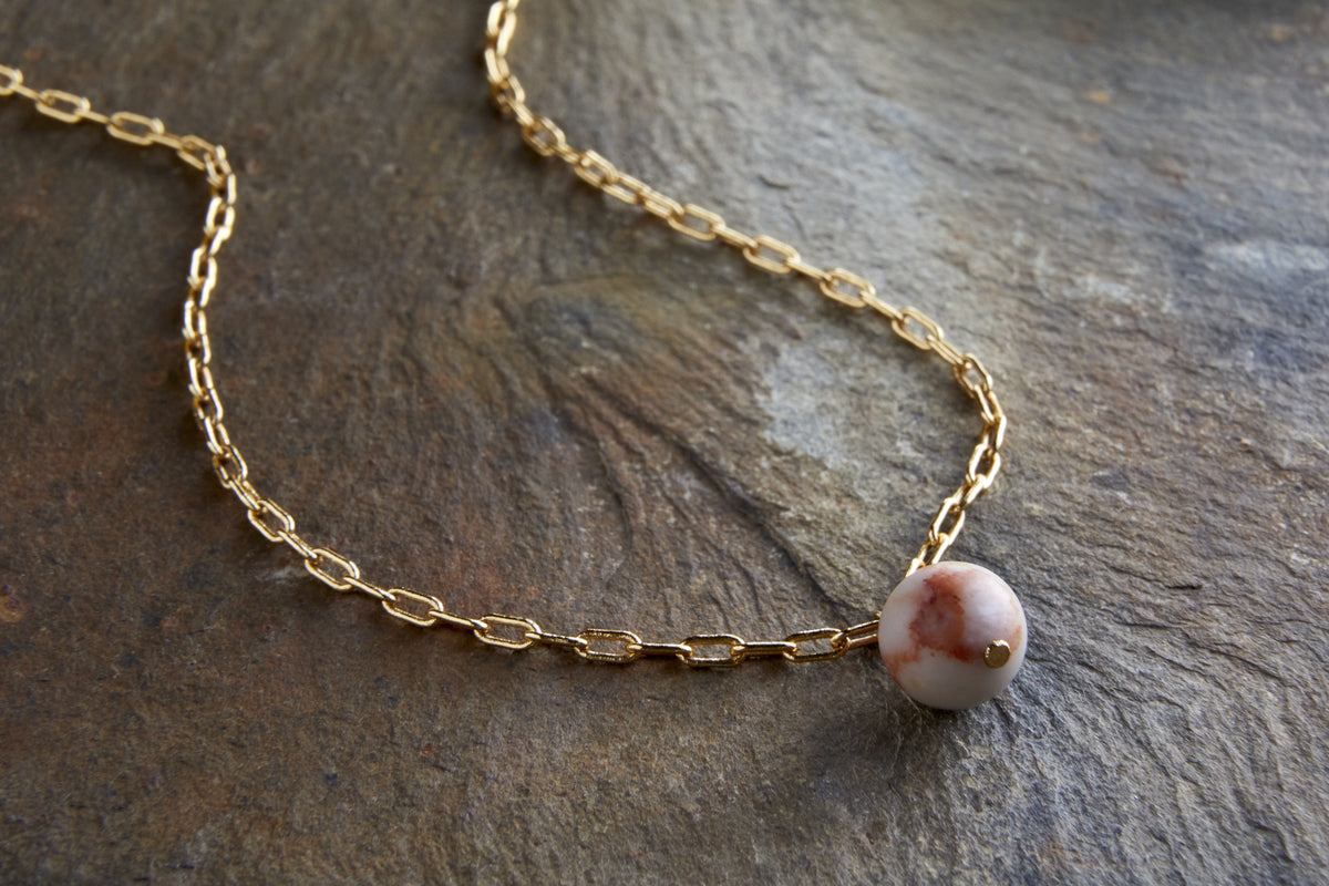 The nkuku Cerita Rhodochrosite Necklace features a gold chain and a round, gold-plated pendant with a marbled white and brown rhodochrosite stone—an elegant jewellery gift for any occasion.