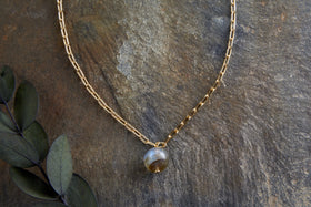 The Marai Labradorite Necklace - Labradorite & Gold by nkuku, featuring elongated gold links and a round marbled labradorite pendant, is displayed on brown stone with eucalyptus leaves—a perfect jewellery gift.
