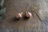 The Cerita Rhodochrosite Stud Earrings by nkuku feature spherical, marbled pink-and-white rhodochrosite stones set in gold-plated settings, displayed on a textured brown stone beside green fabric leaves.