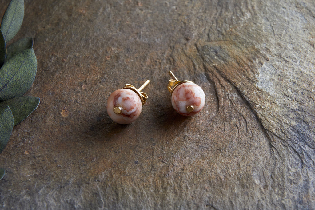 The Cerita Rhodochrosite Stud Earrings by nkuku feature spherical, marbled pink-and-white rhodochrosite stones set in gold-plated settings, displayed on a textured brown stone beside green fabric leaves.