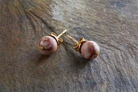 The Cerita Rhodochrosite Stud Earrings by nkuku feature round, marbled pink and white rhodochrosite stones in gold-plated settings, showcased on a textured brown stone surface for an elegant look.