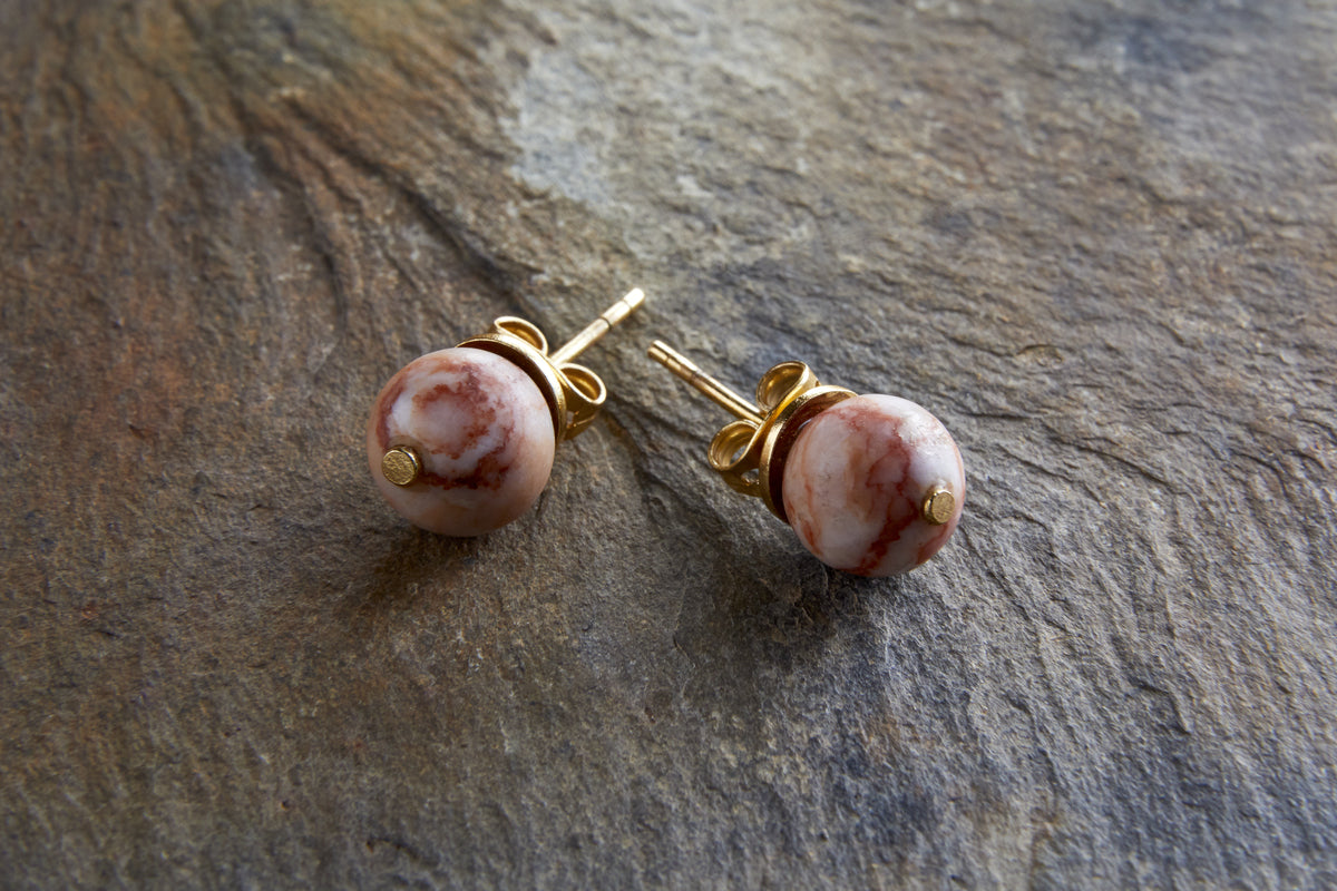 The Cerita Rhodochrosite Stud Earrings by nkuku feature round, marbled pink and white rhodochrosite stones in gold-plated settings, showcased on a textured brown stone surface for an elegant look.