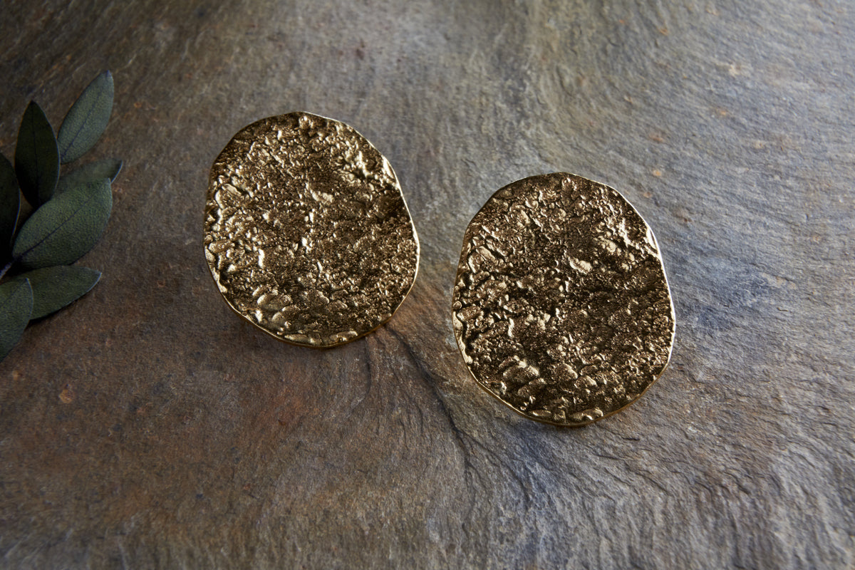 The Oya Hammered Earrings - Gold by nkuku, featuring textured irregular ovals and an artisanal finish, rest on a rough stone surface with green leaves partially visible on the left.