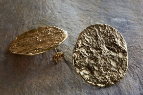 The Oya Hammered Earrings - Gold by nkuku, featuring large, irregular oval shapes and textured finishes, rest elegantly on a stone surface—one flat, the other propped up—highlighting their handcrafted appeal.