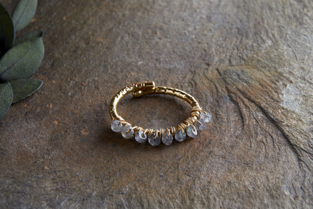 The Vani Labradorite Ring by nkuku, featuring labradorite stones and gold detailing, is coiled in a circle on a textured brown stone surface, with green leaves partially visible in the upper left corner.