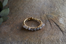The Vani Labradorite Ring by nkuku, featuring labradorite stones and gold detailing, is coiled in a circle on a textured brown stone surface, with green leaves partially visible in the upper left corner.