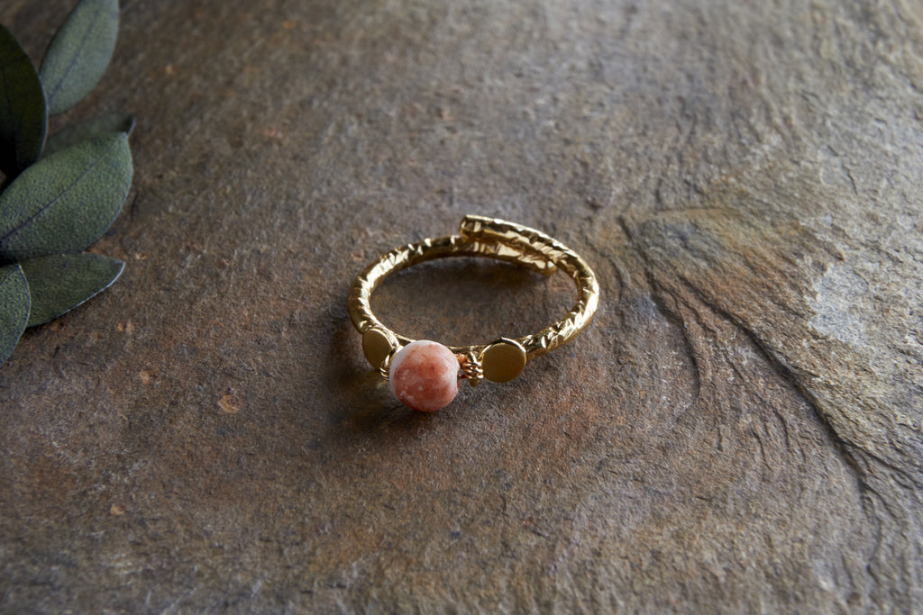 The Cerita Rhodochrosite Ring by nkuku, featuring a pink rhodochrosite stone and textured gold-plated band, is displayed on a rough brown stone with green leaves in the upper left corner.