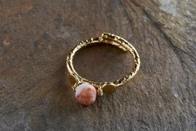 The Cerita Rhodochrosite Ring by nkuku features a twisted, gold-plated band displayed on stone, showcasing a round pinkish-orange rhodochrosite with white marbling, held by wire and flanked by two small gold discs.
