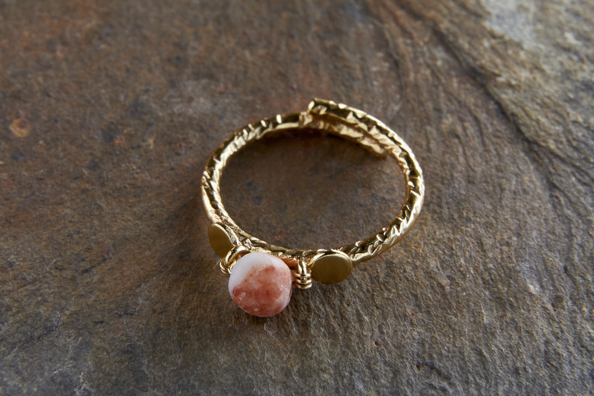 The Cerita Rhodochrosite Ring by nkuku features a twisted, gold-plated band displayed on stone, showcasing a round pinkish-orange rhodochrosite with white marbling, held by wire and flanked by two small gold discs.