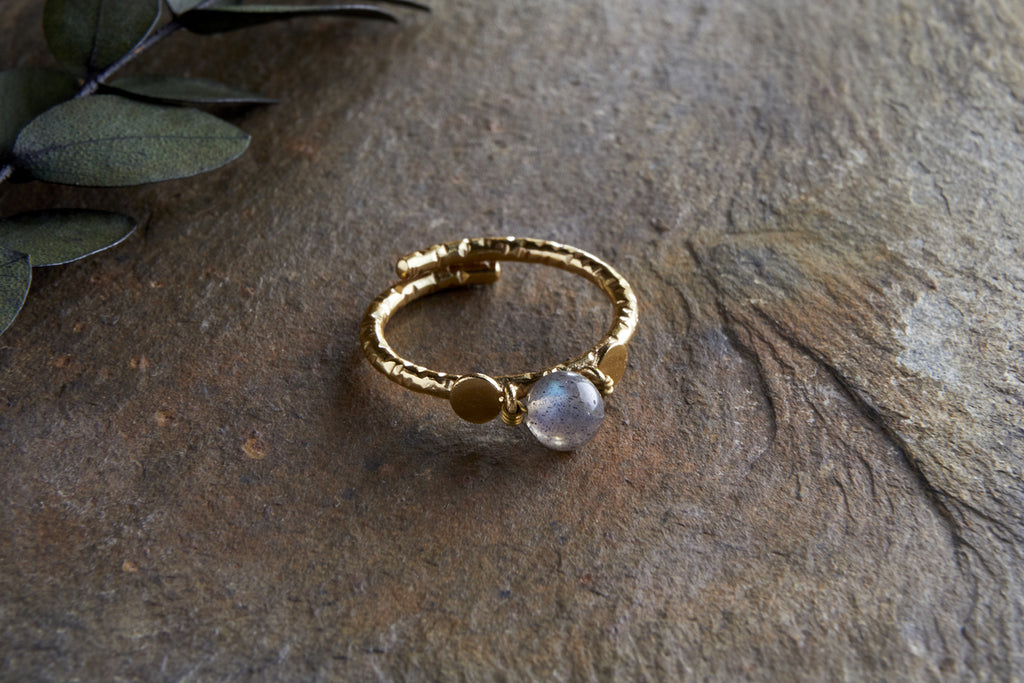The Marai Labradorite Ring by nkuku features a textured gold-plated band with round details and a translucent labradorite centerpiece, displayed on a rough stone surface with green leaves in the upper left corner.