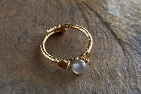 The Marai Labradorite Ring - Labradorite & Gold by nkuku features two flat gold discs and a round, semi-transparent labradorite gemstone with a hammered finish, exuding rustic charm and elegance on a textured stone surface.