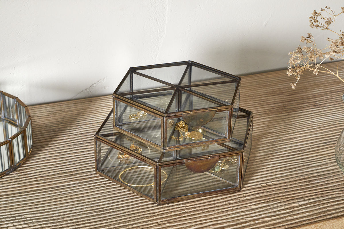 The nkuku Sitara Jewellery Box in antique brass sits on a wooden surface, holding gold jewelry. Nearby are a round mirrored tray and a glass vase with dried flowers, all set against a plain white wall.