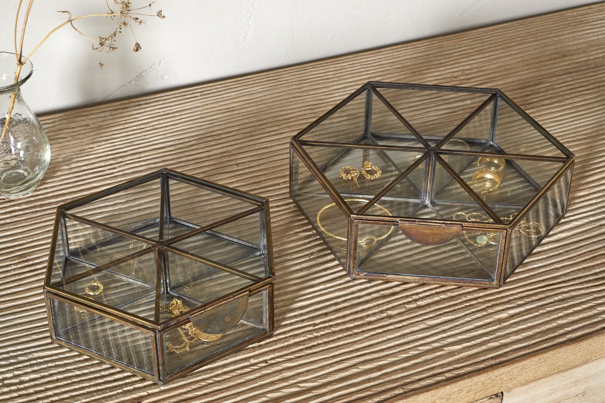 Two nkuku Sitara Jewellery Boxes in antique brass sit on a ribbed wooden surface. The smaller holds a gold bracelet and ring, the larger has earrings, rings, and a necklace. A glass vase with dried flowers is nearby.