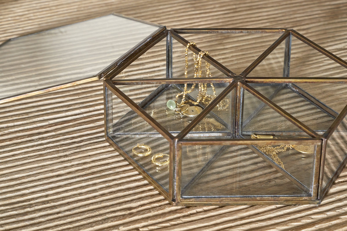 The Sitara Jewellery Box - Antique Brass by nkuku sits open on a ribbed beige surface, displaying gold necklaces, rings, and a green gemstone bracelet neatly inside, their reflections shimmering on the glass sides and base.