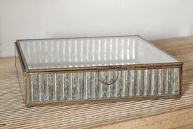 The nkuku Malita Jewellery Box features a gold-toned antique brass frame and ribbed mercury glass sides with a small metal ring pull, displayed on a light wood surface against a white wall.
