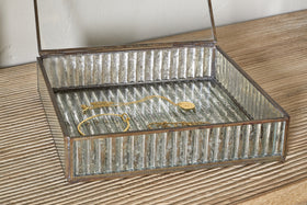 The Malita Jewellery Box by nkuku, featuring vertical ridged glass and an antique brass frame, sits open on light wood. A gold necklace and bracelet rest neatly inside on the ribbed glass beneath the hinged lid.