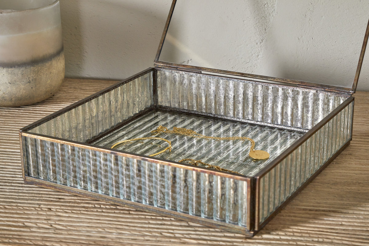 The nkuku Malita Jewellery Box in antique brass, featuring a ribbed rectangular design and hinged lid, sits open on a wooden surface with a gold bracelet and necklace inside. A frosted mercury glass candle holder stands to its left.
