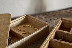The nkuku Chanya Mango Wood Jewellery Box - Natural, with compartments for treasures, sits open on a rustic surface displaying a gold chain pendant, its natural wood grain set against a softly textured wall.