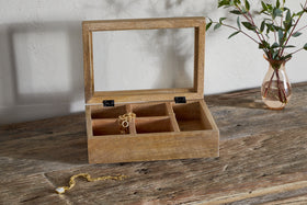 The nkuku Kabani Mango Wood Jewellery Box - Natural, featuring an open glass lid and four compartments with gold jewellery, sits on a rustic table. A gold bracelet with a pearl charm is outside, and a glass vase with leafy branches is behind.