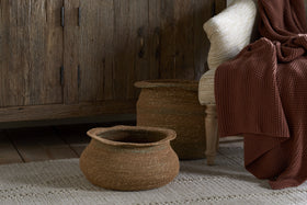 The nkuku Rana Short Baskets in Natural & Moss sit on a textured beige rug next to a wooden cabinet, while a light chair with a chunky rust-red knit blanket adds to the cozy, rustic look—ideal for fans of natural home decor.