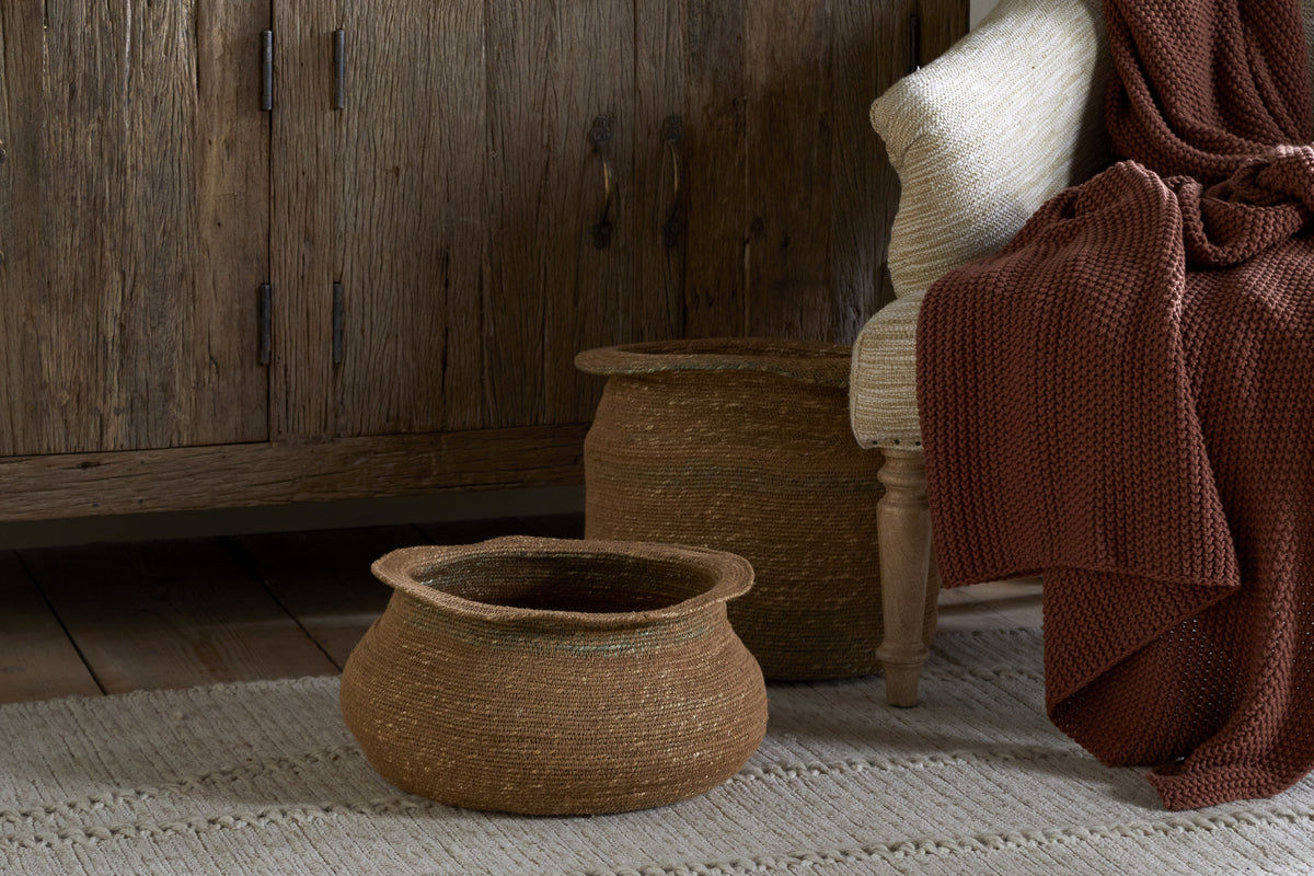 The nkuku Rana Short Baskets in Natural & Moss sit on a textured beige rug next to a wooden cabinet, while a light chair with a chunky rust-red knit blanket adds to the cozy, rustic look—ideal for fans of natural home decor.