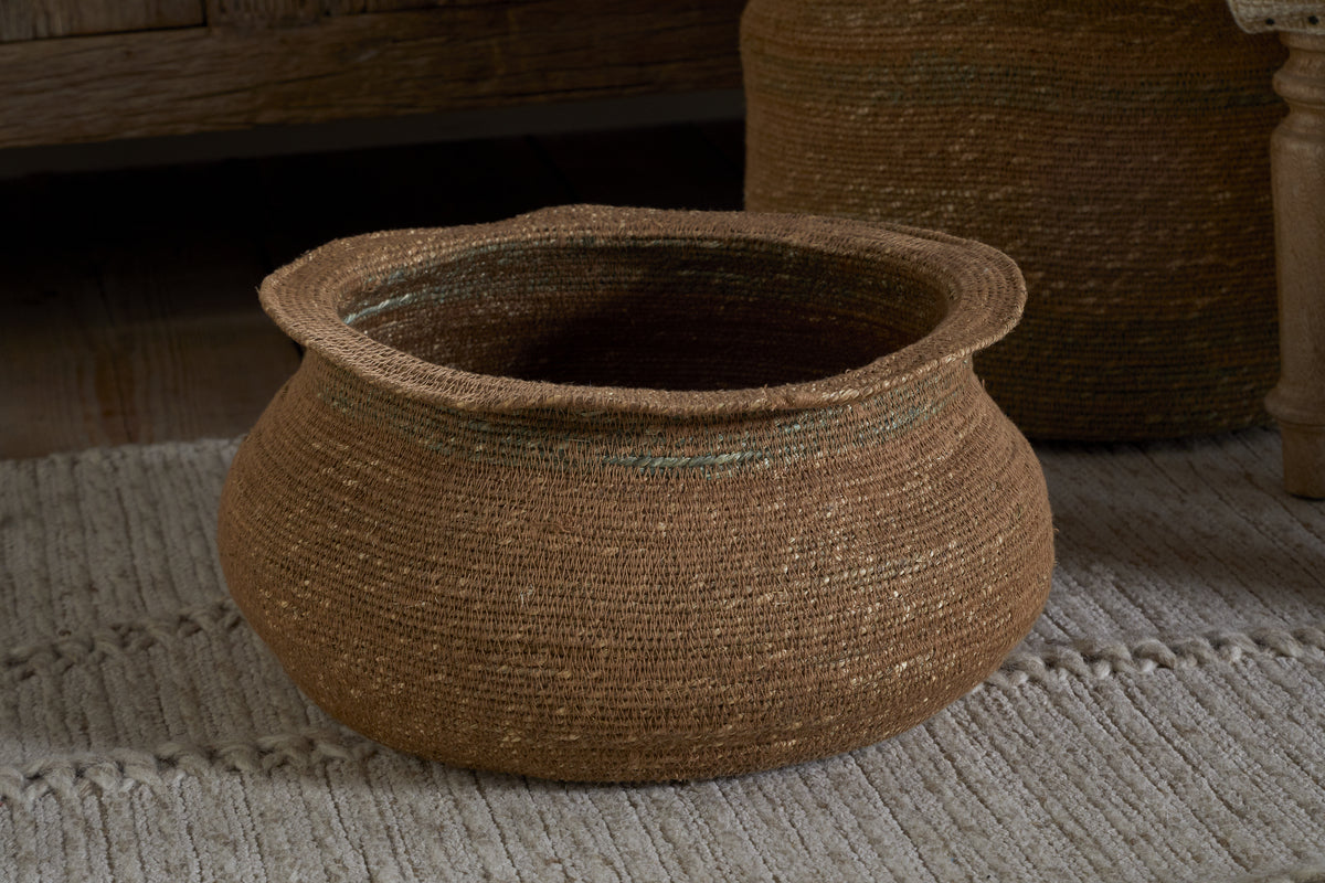 The nkuku Rana Short Basket - Natural & Moss features a handwoven round seagrass design with visible weave patterns and moss-green accents—ideal for natural home decor and pictured here on a textured, off-white rug.