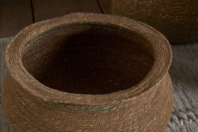 A close-up of the nkuku Rana Short Basket in Natural & Moss, crafted from natural brown fibers with a wide opening, highlights its detailed weaving. Perfect for storage or as decor, it sits on a beige rug with another similar basket in the background.