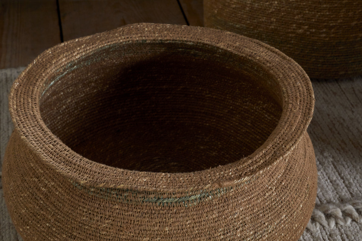 A close-up of the nkuku Rana Short Basket in Natural & Moss, crafted from natural brown fibers with a wide opening, highlights its detailed weaving. Perfect for storage or as decor, it sits on a beige rug with another similar basket in the background.