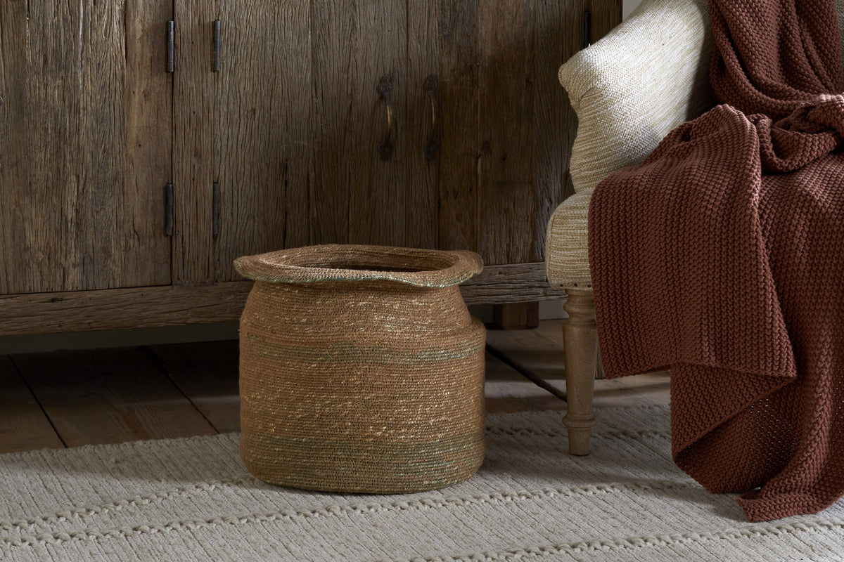 The nkuku Rana Tall Basket in Natural & Moss sits on a textured rug by a rustic wooden cabinet. Nearby, a wooden chair with a cream cushion and maroon-brown knit blanket adds to the cozy, earthy vibe.