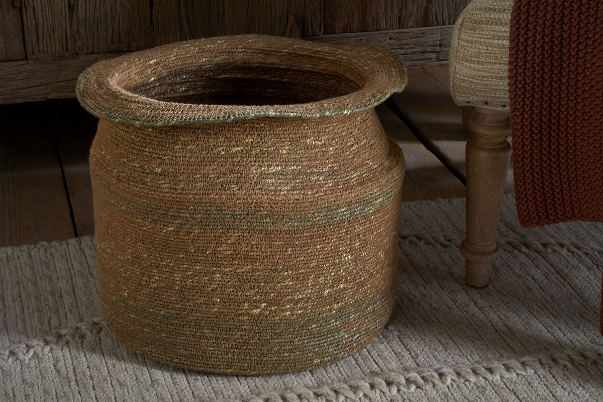 The nkuku Rana Tall Basket - Natural & Moss, a large round seagrass basket in brown and tan, sits on a textured beige rug. With its wide mouth and flared rim, it offers stylish storage. A wooden chair with woven upholstery is to the right.