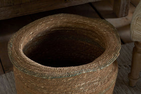 The nkuku Rana Tall Basket in Natural & Moss, featuring an uneven rim, sits empty on a textured light rug near wooden furniture and an upholstered chair.