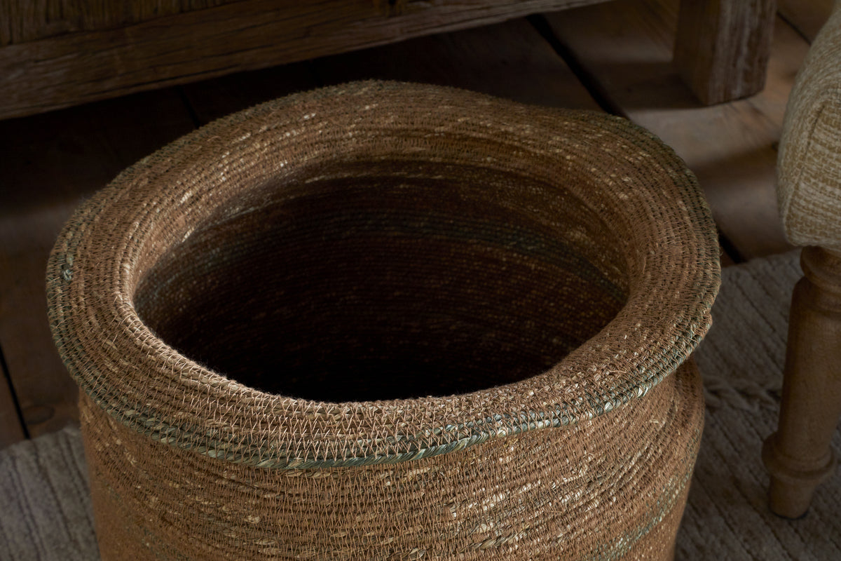 The nkuku Rana Tall Basket in Natural & Moss, featuring an uneven rim, sits empty on a textured light rug near wooden furniture and an upholstered chair.