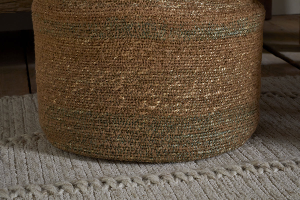 The nkuku Rana Tall Basket - Natural & Moss features subtle greenish threads and sits on a light gray, textured rug with raised lines, adding decorative storage atop wood flooring.