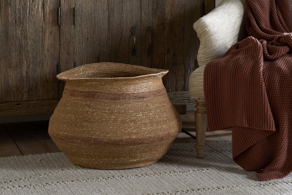 The nkuku Rana Wide Basket in Natural & Rust sits on a textured rug by a wooden cabinet, while a beige chair nearby is draped with a rust knit blanket, creating a cozy and rustic atmosphere.