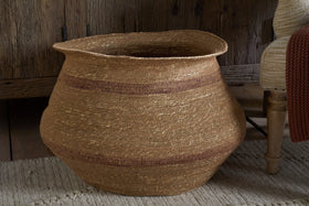 The nkuku Rana Wide Basket in Natural & Rust, featuring handwoven seagrass with subtle brown stripes, sits on a beige textured rug beside a wooden cabinet and upholstered chair, adding rustic warmth to the space.