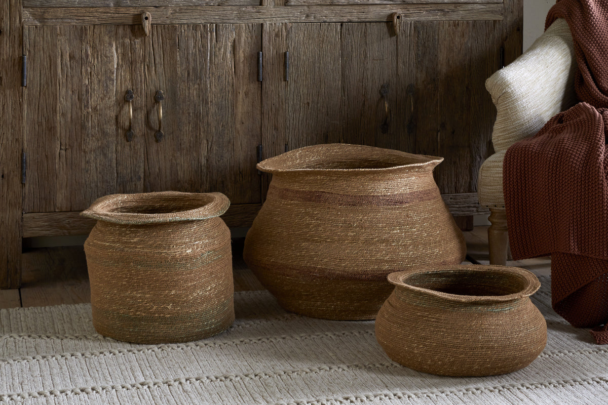 Three Rana Wide Baskets by nkuku in natural and rust tones are arranged on a cream rug before a rustic cabinet. A cream chair with a rust knit blanket adds warmth to this inviting storage basket display.