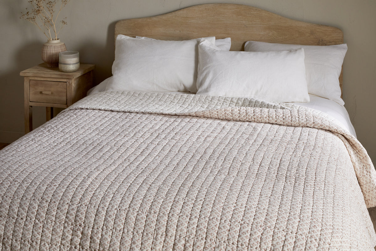 A neatly made bed features the nkuku Peplu Cotton & Linen Screen Print Bed Quilt in rust, three white pillows, and a wooden headboard. A wooden nightstand with a vase of dried flowers and decor accents stands by a neutral wall.