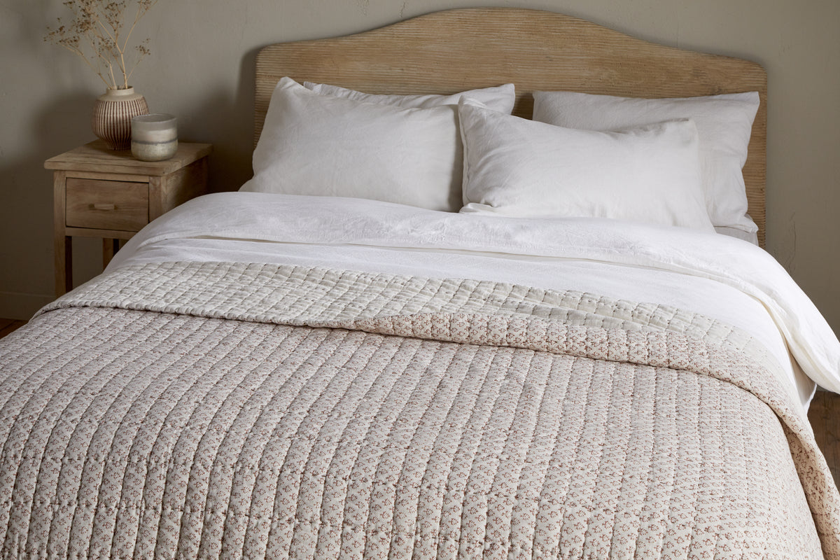 A neatly made bed with a light wood headboard, four white pillows, and the Peplu Cotton & Linen Screen Print Bed Quilt in rust by nkuku. A wooden nightstand with a vase and small decor sits to the left against a neutral wall.