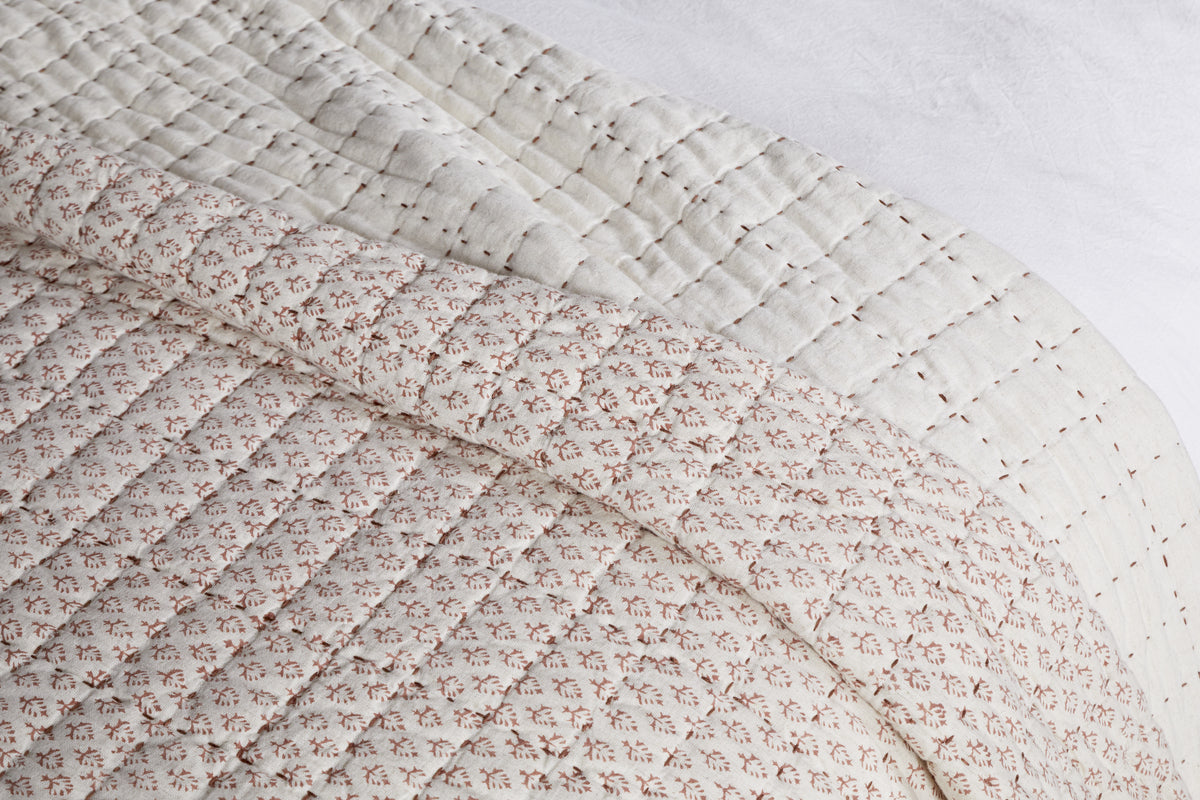 A close-up of nkuku's Peplu Cotton & Linen Screen Print Bed Quilt - Rust draped over a bed highlights its soft, textured finish with a rust-toned pattern on top and an off-white underside featuring subtle stitched lines.