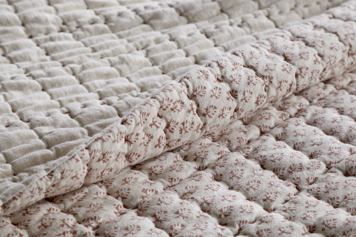 A close-up of the nkuku Peplu Cotton & Linen Screen Print Bed Quilt in Rust shows a folded corner, highlighting its soft, textured surface and intricate repeating pattern—showcasing handmade Indian artistry and cozy appeal.