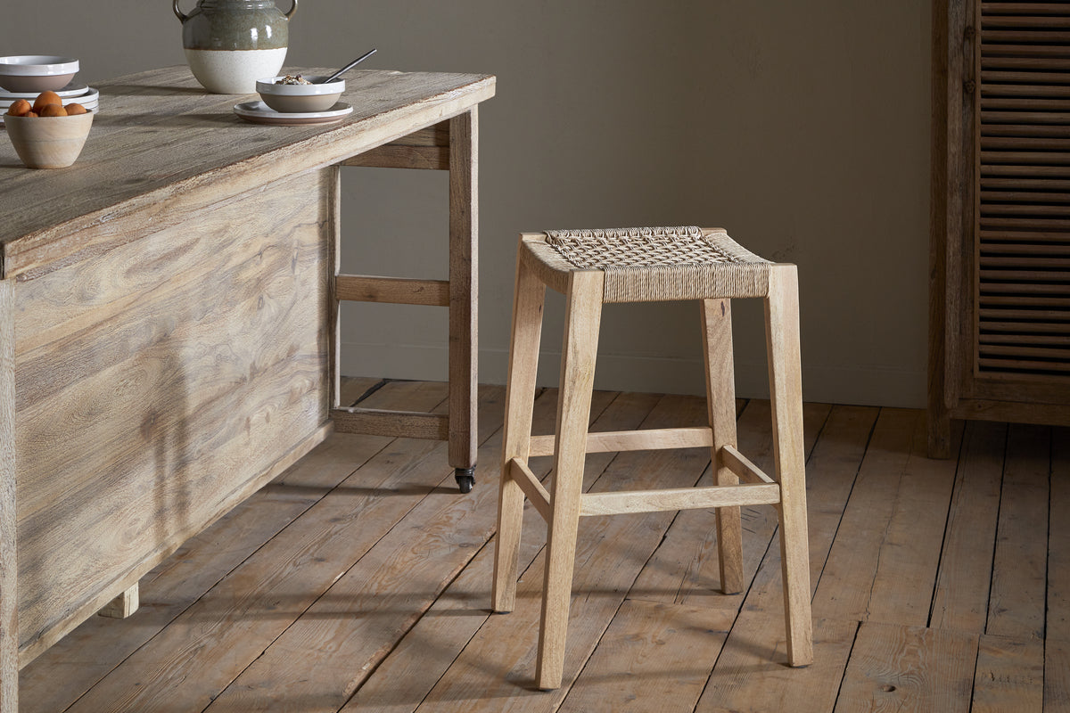 A natural wood kitchen with a rustic breakfast table, set with bowls and a jug, is complemented by the nkuku Aamani Mango Wood & Cord Counter Stool - Natural, featuring a woven seat. Wooden floors and soft neutral lighting complete the scene.