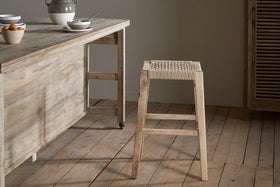 A rustic kitchen shows a light mango wood table set for breakfast and the Aamani Mango Wood & Cord Counter Stool by nkuku, featuring a woven seat in natural tones. The wide plank floor and neutral backdrop complete the scene.