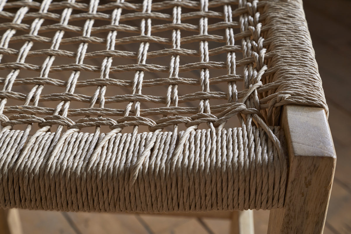 Close-up of the nkuku Aamani Mango Wood & Cord Counter Stool - Natural, featuring a thick, natural-colored rope seat woven in a crisscross pattern on a light mango wood frame. The detailed craftsmanship and texture are clearly visible. Wooden floor background.