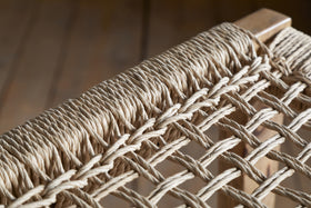Close-up of the nkuku Aamani Mango Wood & Cord Counter Stool - Natural, featuring a tightly woven natural-colored rope seat on a smooth mango wood frame. Blurred wooden flooring appears in the background.